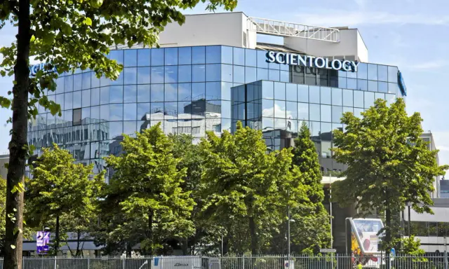 Bâtiment moderne aux façades vitrées reflétant le ciel bleu, avec le nom "SCIENTOLOGY" inscrit en lettres bleues lumineuses. Des arbres verdoyants entourent la structure.