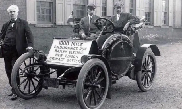 Un hombre junto a un coche eléctrico Bailey de 1907, parte de la historia del coche eléctrico, con un cartel que anuncia una carrera de resistencia de 1000 millas.