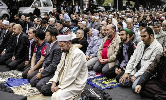 Männer in traditioneller Kleidung knien auf Gebetsteppichen. Ein Mann in einem weißen Gewand und Turban leitet das Gebet, ein Zeichen der berberischen Religion.