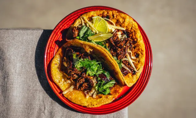 Zwei saftige Birria Tacos auf einem roten Teller, garniert mit Koriander und Limettenspalten.