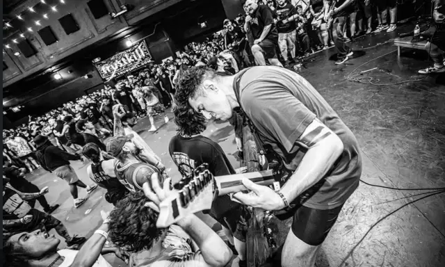 Energetyczny koncert w klubie. Czarno białe zdj. z tłumem szalejącym pod sceną, gdzie gitarzysta pochyla się nad instrumentem.