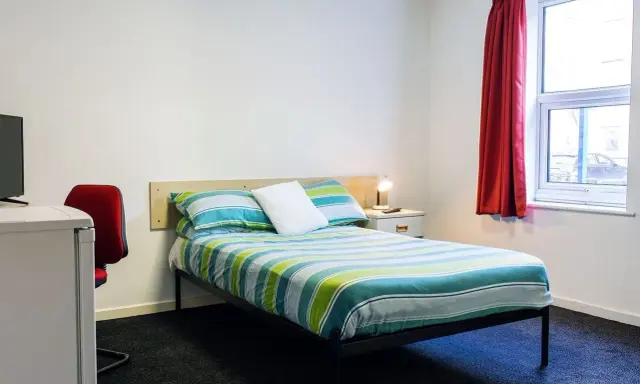 Habitación en Atlantic Point, Liverpool, con cama a rayas verdes y azules, escritorio rojo y ventana con cortina roja.