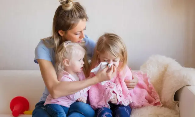 Mama wyciera nos chorej córce, podczas gdy młodsza siostra patrzy. Czy szczepienie mogłoby zapobiec katarowi?