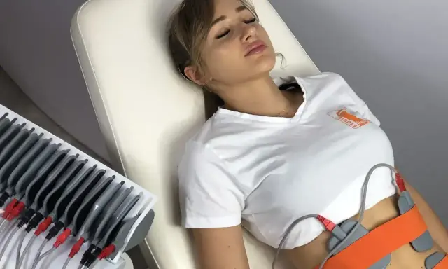 Kobieta leży na kozetce, poddana zabiegowi elektrostymulacji brzucha. Elektrody przymocowane są do jej talii.