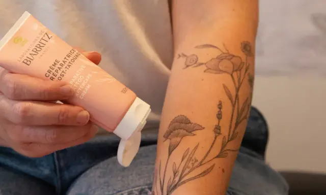 Une main tient un tube de crème réparatrice post-tatouage, prêt à être appliqué sur un bras orné d'un tatouage floral délicat.