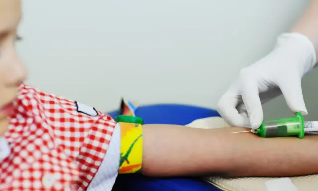 Dziecko w czerwono-białą kratkę ma pobieraną krew. Lekarz w białej rękawiczce pobiera próbkę do analizy, która może wykazać, czy pct podwyższone u dziecka.