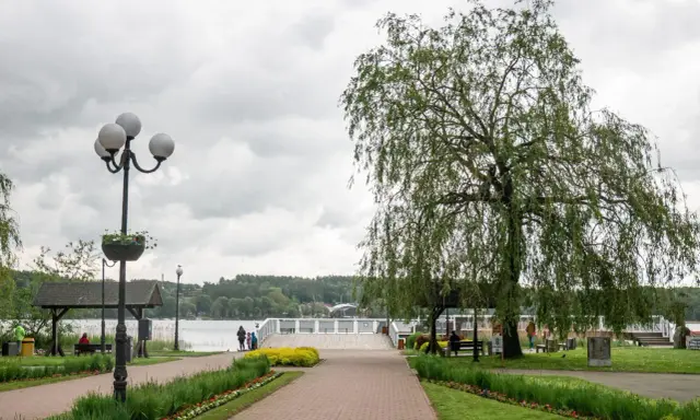 Spacer w Mrągowie nad jeziorem, z wierzbą i kwitnącymi rabatami. Idealne miejsce na relaks i podziwianie mrągowskich atrakcji.