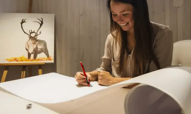 En kvinna ler medan hon ritar på ett stort papper. I bakgrunden syns en oljemålning av en ren. Hon deltar i en oljemålning kurs.