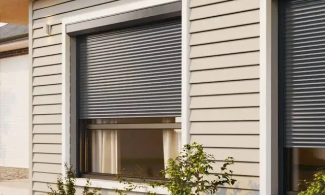 Volet roulant gris fermé sur une fenêtre de maison avec bardage beige.
