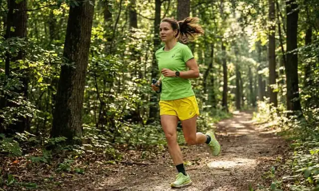 Frau joggt durch einen sonnendurchfluteten Wald. Sie trägt ein grünes Shirt und gelbe Shorts. Das zeigt, was bringt joggen: Natur, Bewegung und Wohlbefinden.