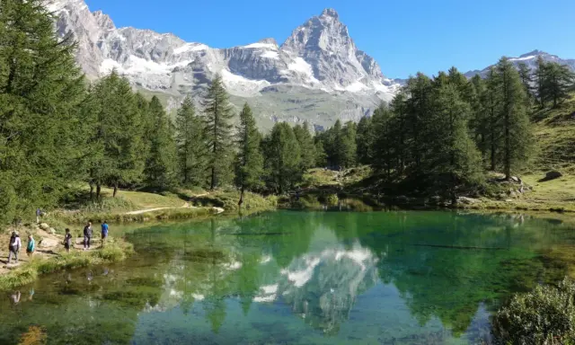 Un gruppo di escursionisti ammira il riflesso del Cervino nel lago blu di Cervinia, circondato da pini e montagne innevate.