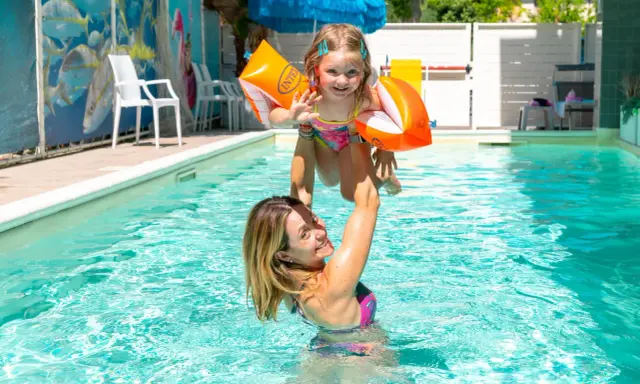 Mamma solleva la figlia con braccioli arancioni in una piscina, un momento di gioia in un bagno giardino a Cesenatico.