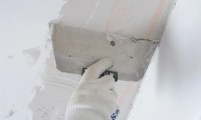 Ręka w rękawicy nakłada zaprawę na styropian przy suficie. Proces pokazujący, jak przykleić styropian do sufitu.