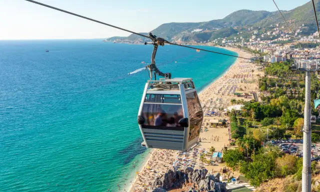 Seilbahn über dem belebten Alanya Strand mit türkisfarbenem Meer und vielen Sonnenanbetern.