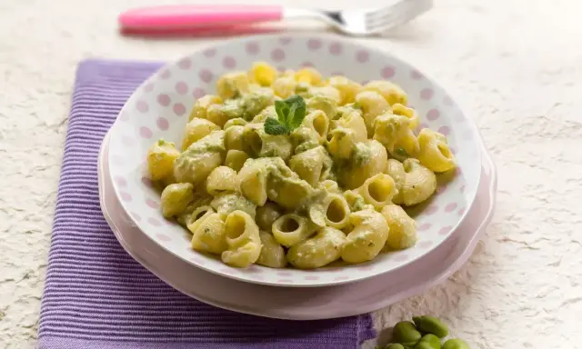 Piatto di pasta con le fave, condita con una crema verde e guarnita con menta. Un contorno di fave fresche completa la scena.