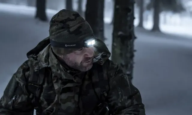 Mężczyzna w zimowej, kamuflującej kurtce i czapce z czołówką Olight, gotowy na wyprawę w góry.