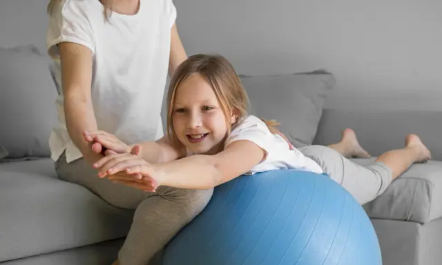 Dziecko z obniżonym napięciem mięśniowym ćwiczy mowę, leżąc na piłce rehabilitacyjnej pod opieką terapeuty.