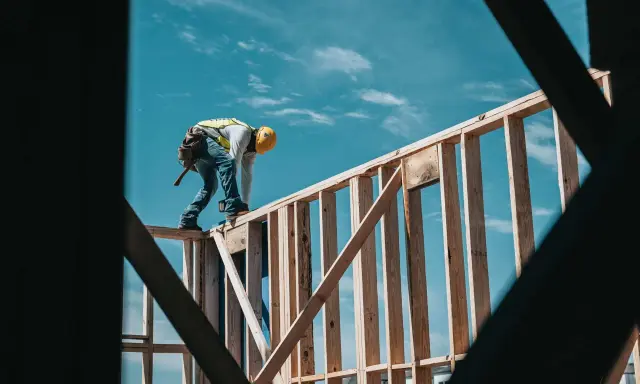 Obrero con casco amarillo y chaleco reflectante trabaja en la estructura de madera de una casa. El cielo azul con nubes es el fondo. El motor de su moto es de 1544 cc.