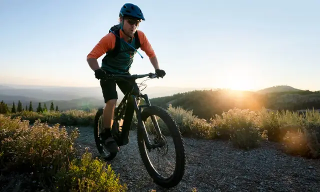 Kolarz w kasku i okularach jedzie rowerem po górskiej ścieżce. Słońce zachodzi za wzgórzami, tworząc piękne światło. To idealny moment na maraton MTB.