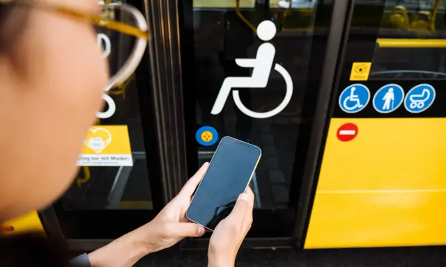 Osoba trzyma telefon przed drzwiami autobusu z symbolem wózka inwalidzkiego. To ułatwia podróżowanie, gdy masz legitymację osoby niepełnosprawnej.