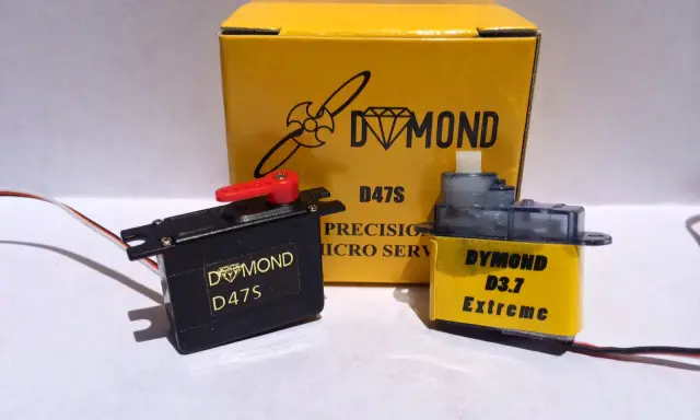 Zwei Servos, ein schwarzer Dymond D47S und ein gelber Dymond D3.7 Extreme, stehen vor einer gelben Box mit der Aufschrift "Dymond D47S".