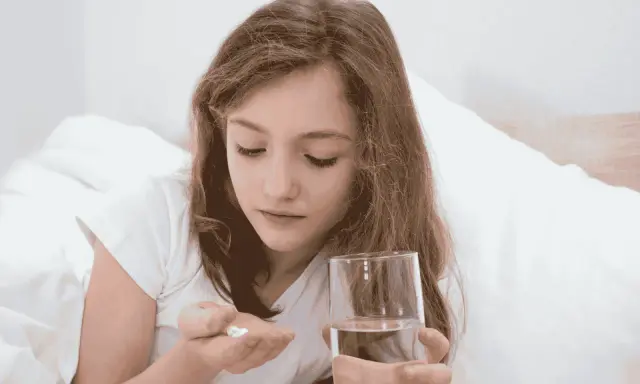 Dziewczynka w łóżku trzyma w dłoni tabletki i szklankę wody. Uważnie patrzy na białe tabletki, które mogą być przeterminowany paracetamol dla dzieci.