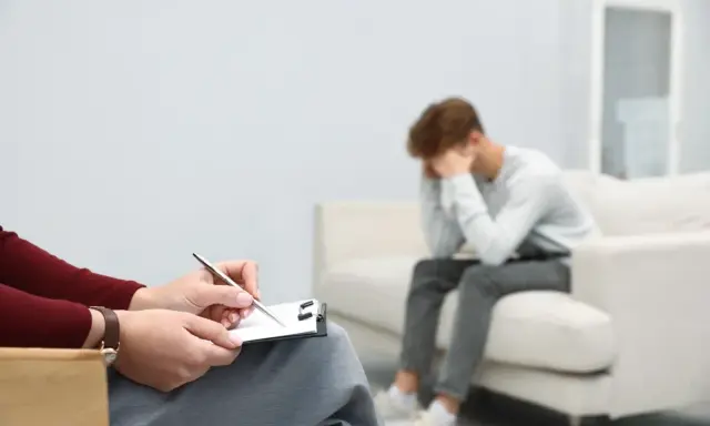 Młody mężczyzna siedzi na kanapie, ukrywając twarz w dłoniach, przeżywając cierpienie psychiczne. Obok niego terapeuta notuje coś na kartce.