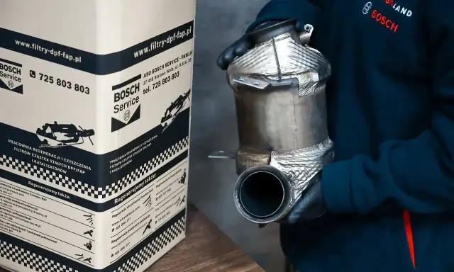 Osoba w rękawiczkach trzyma filtr DPF/FAP. Obok stoi karton z napisem "www.filtry-dpf-fap.pl", sugerującym, że to miejsce zajmuje się regeneracją.