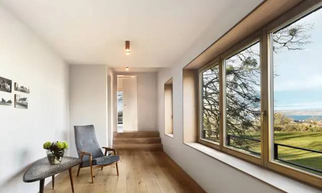 Interior moderno con suelo de madera, sillón gris y gran ventanal. El marco de madera de la ventana, que parece recién restaurado, enmarca un paisaje exterior.
