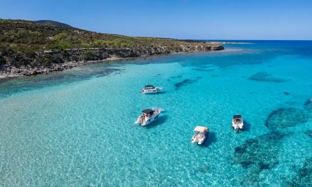 Traumhafter zypern strand mit kristallklarem Wasser und vier Booten, die vor der felsigen Küste ankern.