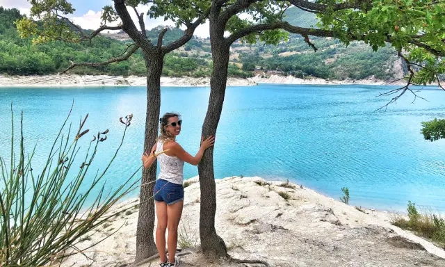Donna sorridente abbraccia un albero con un lago turchese sullo sfondo. Una perfetta gita fuori porta nelle Marche.