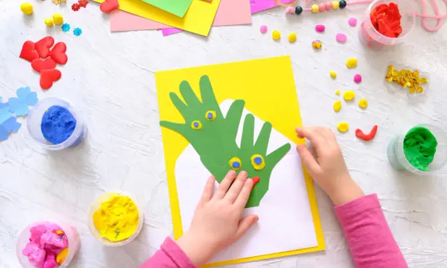 Kreatives Basteln mit Kleinkindern: Hände werden zu grünen Drachen auf gelbem Papier, umgeben von bunter Knete und Bastelmaterialien.