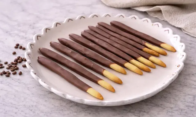 Assiette de mikado maison, bâtonnets de biscuit enrobés de chocolat, avec quelques grains de café à côté.