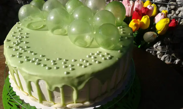 Zielony tort udekorowany koralikami i przezroczystymi, zielonymi balonami z żelatyny. Obok bukiet kolorowych tulipanów.