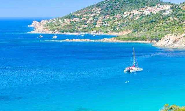Un catamaran blanc avec des accents rouges navigue sur des eaux bleues cristallines, près d'une côte verdoyante et d'un village pittoresque.