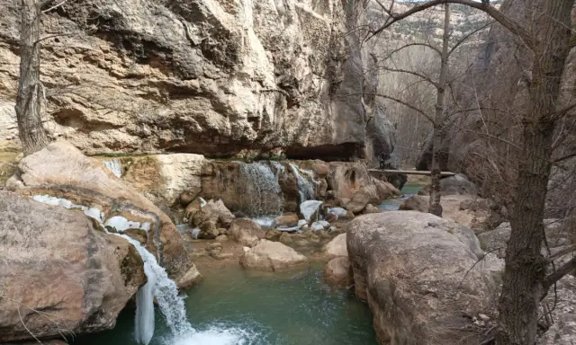 Cascada y río en un cañón rocoso, con hielo en las rocas. Un puente de madera cruza el río, evocando las pasarelas de Valloré.