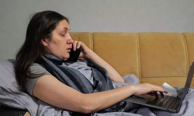 Kobieta w łóżku, rozmawiająca przez telefon i pracująca na laptopie, zastanawia się, na ile dni opłaca się brać zwolnienie lekarskie.