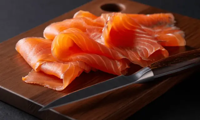 Finas lonchas de salmón ahumado, listas para disfrutar, reposan sobre una tabla de madera junto a un cuchillo afilado.
