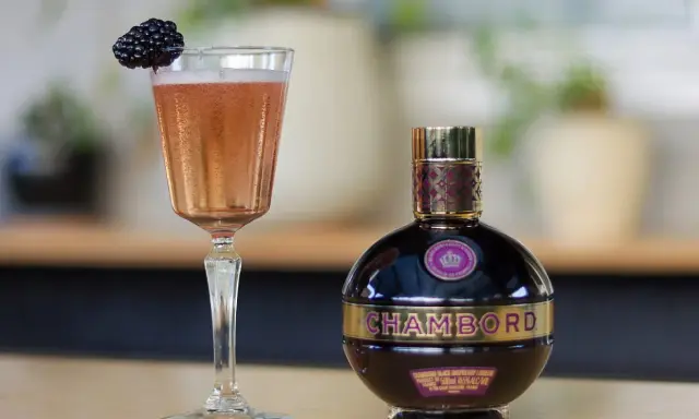 Elegancki kieliszek z różowym drinkiem i jeżyną obok butelki likieru Chambord.