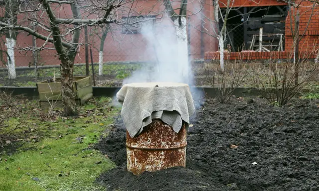 Dym unosi się z beczki, która służy jako prowizoryczna wędzarnia. Wokół niej widać ziemię i trawę.