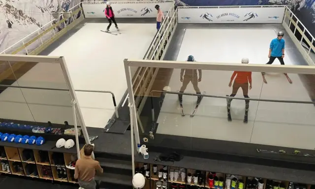 Ludzie ćwiczą jazdę na nartach na sztucznym stoku narciarskim w Ski Arenie Wrocław. Widać sprzęt narciarski i buty narciarskie w schowkach.
