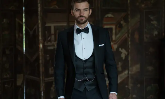 A dapper man in a black suit wedding guest attire, complete with a bow tie and waistcoat, stands confidently.