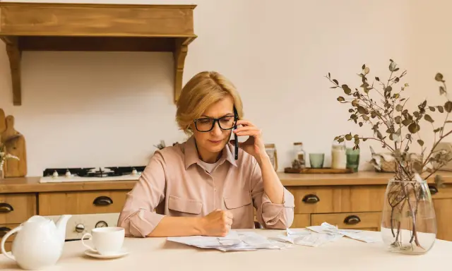 Kobieta w okularach rozmawia przez telefon, siedząc przy stole w kuchni. Przed nią rachunki. Prowadzi **osobne gospodarstwo domowe z mężem**, organizując finanse.