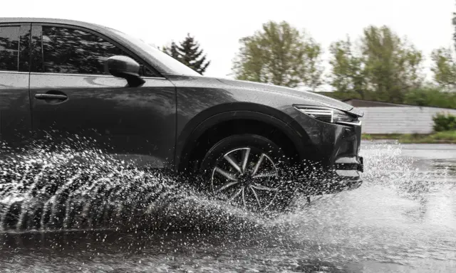 Ciemny samochód wpada w kałużę, powodując efekt aquaplaningu. Woda rozpryskuje się na boki, tworząc białą mgiełkę.