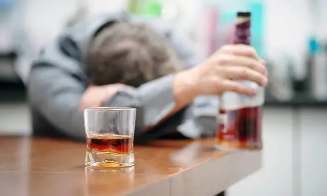 Zaniedbany mężczyzna, prawdopodobnie po eksmisji alkoholika z domu, leży na stole obok szklanki z whisky i butelki.
