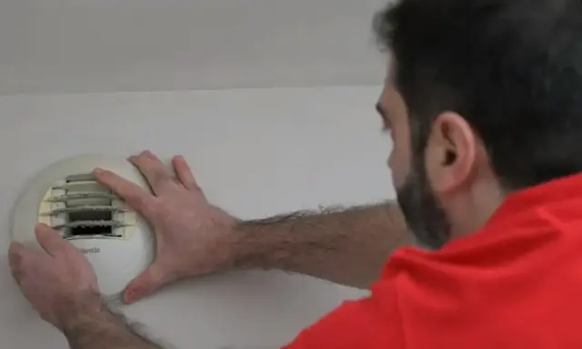 Un homme en chemise rouge effectue l'entretien d'une VMC murale, vérifiant le filtre encrassé.