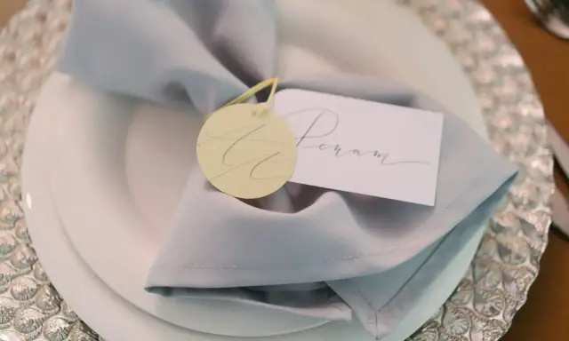Elegancka winietka z imieniem "Peram" na jasnoszarym obrusie, ozdobiona okrągłym, żółtym tagiem.