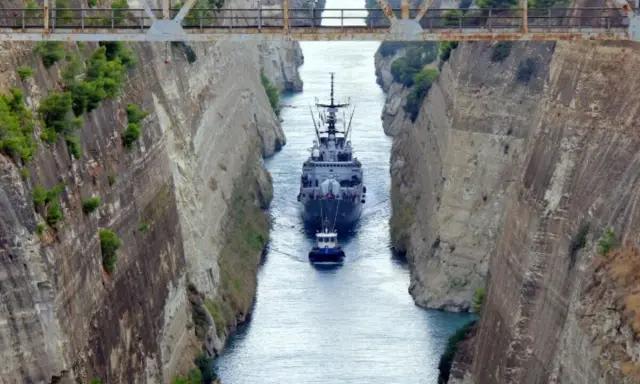 Una nave militare e un rimorchiatore navigano nel **Canale di Corinto**, tra alte pareti rocciose scavate dall'uomo.