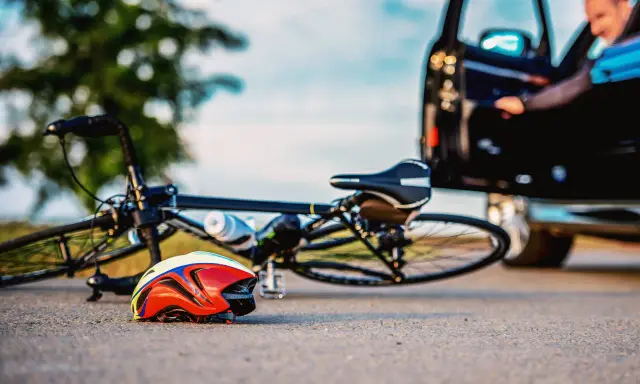 Rower leży na drodze obok kasku. Kierowca wychodzi z czarnego samochodu, sugerując potrącenie rowerzysty bez wezwania policji.