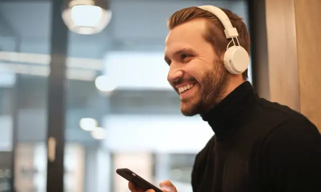Uśmiechnięty mężczyzna w białych słuchawkach na uszach trzyma telefon, być może właśnie uczy się, jak sparować słuchawki z telefonem.